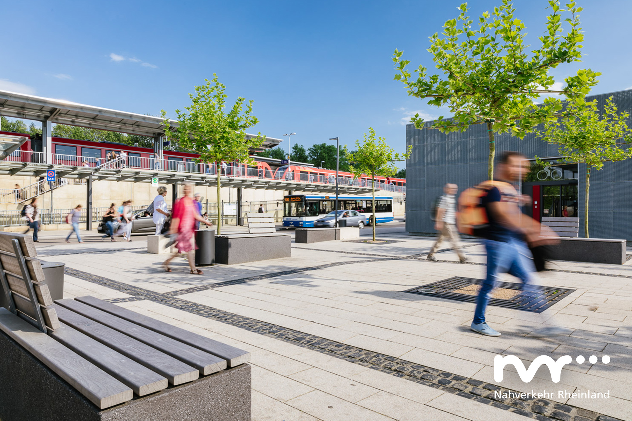 Das Bild zeigt eine sommerliche Impression der Mobilstation in Erftstadt. Mehrere Fahrgäste gehen über die weitläufige Anlage.