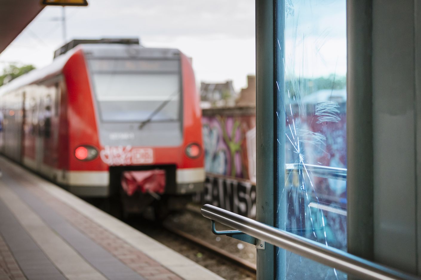 An einem Treppenaufgang zum Gleis ist zerstörtes Glas zu sehen. Im Hintergrund erkannt man zahlreiche Grafittis an Bahn und Außenanlagen.